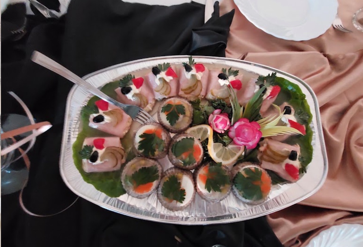 Dekoracyjna taca z przekąskami: roladki z szynki z dodatkami i galaretki z jajkiem i natką pietruszki, ozdobiona rzeźbionymi kwiatami z warzyw i cytryną, na stole z czarno-złotym obrusem.