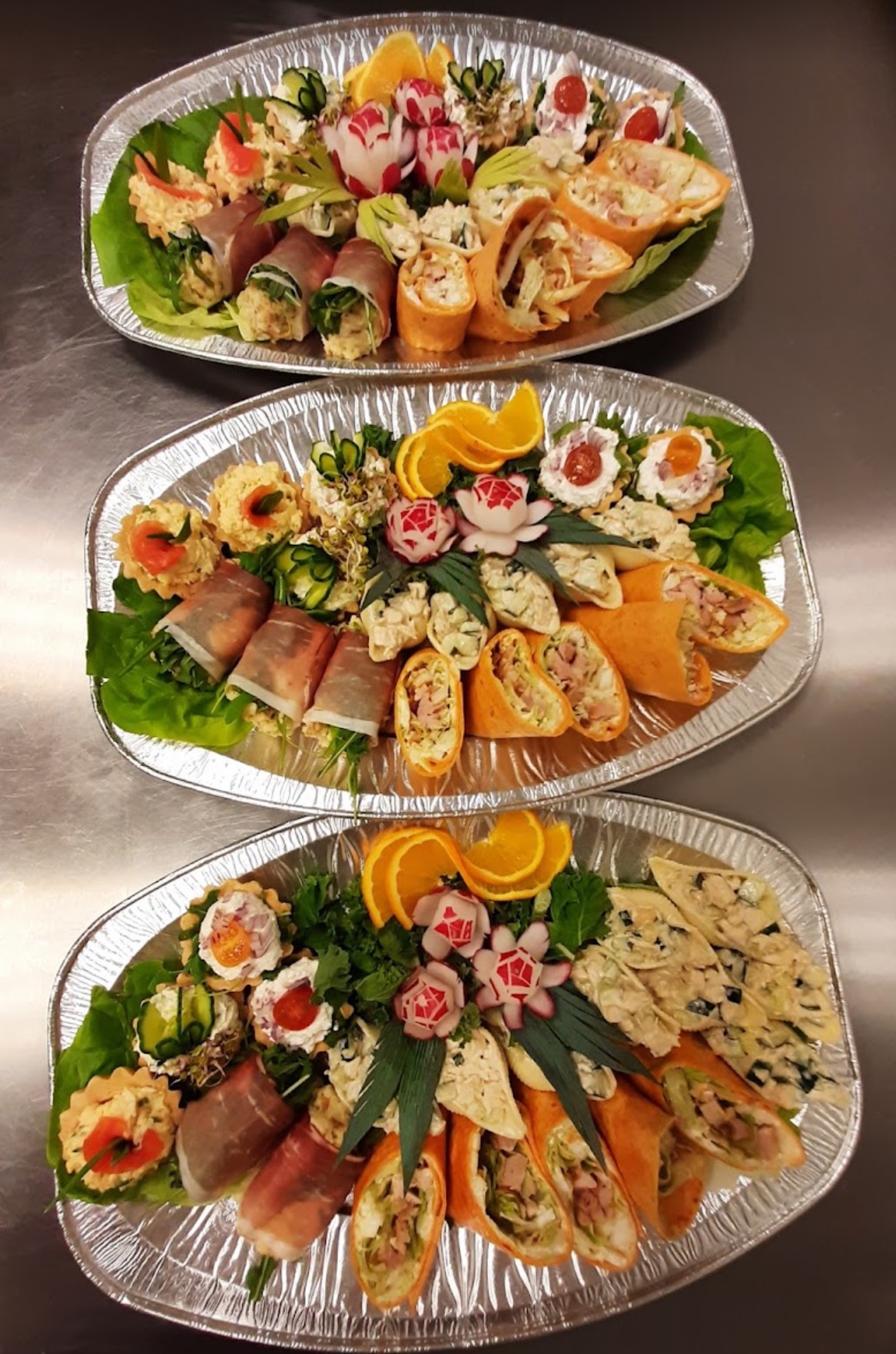 Trzy srebrne tace z różnorodnymi przekąskami: roladki z szynki, tortille z farszem, tartaletki z kremem, dekoracje z rzodkiewki i plastry pomarańczy, ułożone na zielonych liściach sałaty.
