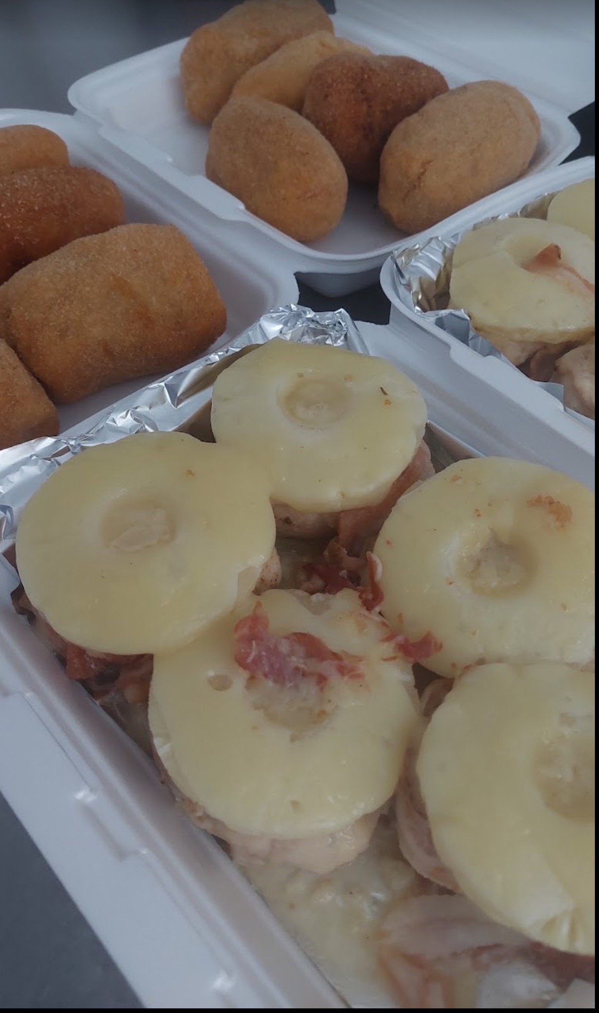 Dwa białe pojemniki styropianowe z przekąskami: krokiety w panierce i danie z plastrami ananasa, szynki i sera, całość na białym sosie.