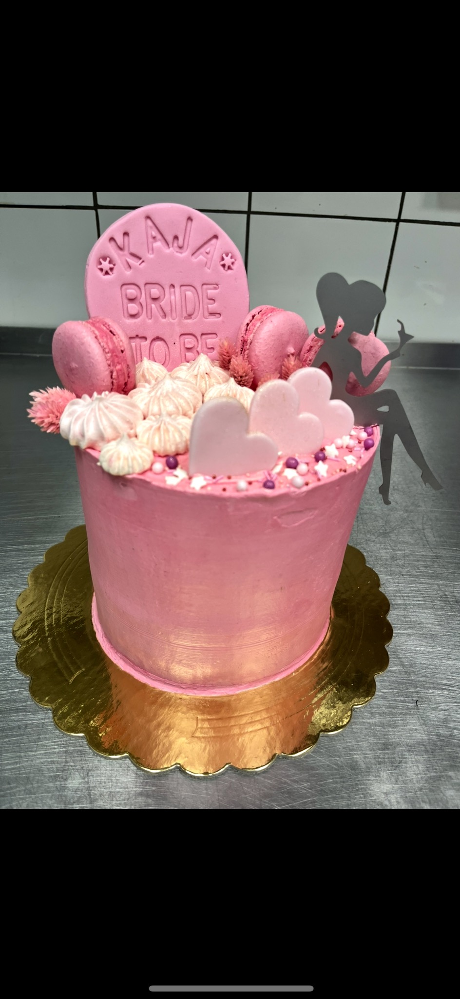 Różowy tort na złotym podkładzie, udekorowany makaronikami, bezami, różowymi sercami i topperem z napisem 'Kaja Bride to be' oraz sylwetką kobiety.