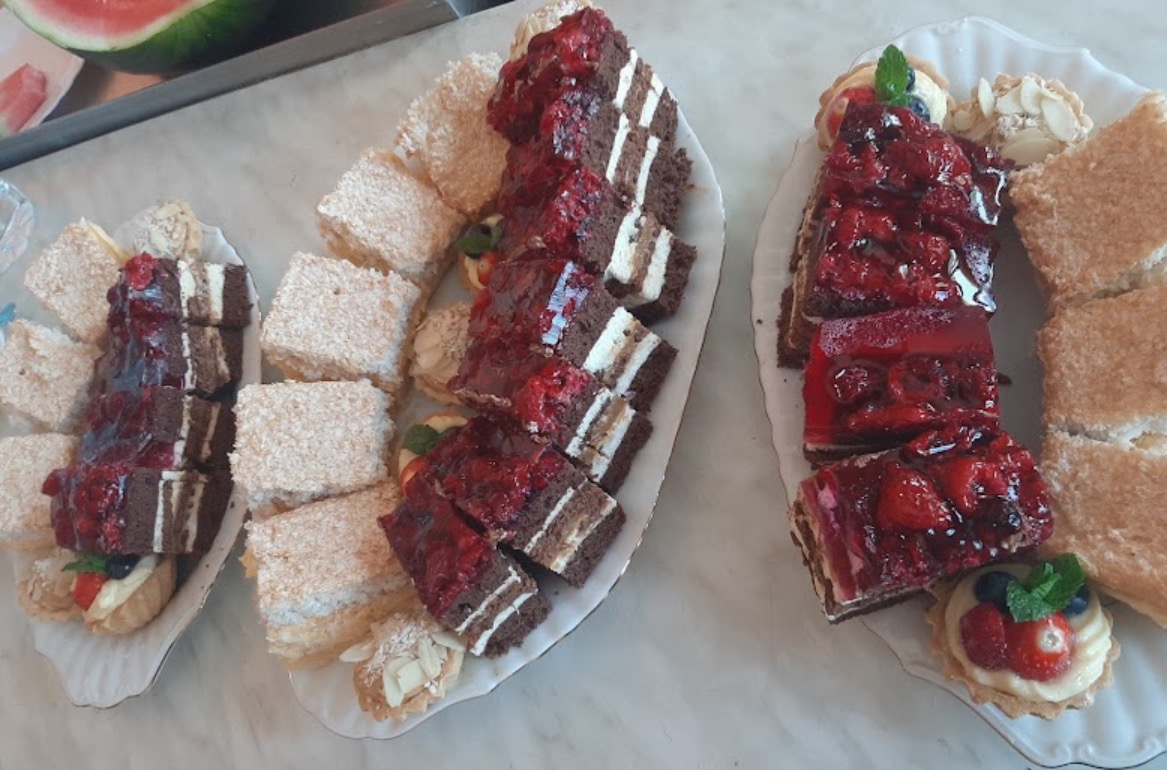 Dwa podłużne talerze z różnorodnymi ciastami: czekoladowe z wiśniami, kokosowe kwadraty, kruche babeczki z kremem i owocami, oraz kawałek arbuza w tle.