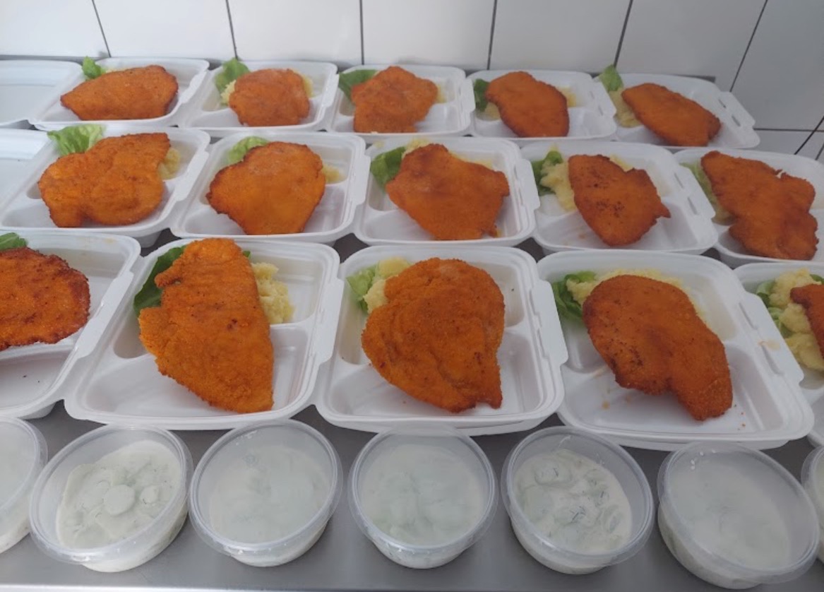 Przygotowane porcje obiadowe z kotletem schabowym, ziemniakami i sałatą w białych pojemnikach cateringowych, uzupełnione o sos w plastikowych kubeczkach, gotowe do transportu w Bydgoszczy.