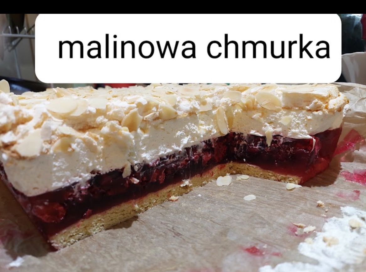 Kawałek ciasta 'Malinowa Chmurka' z biszkoptowym spodem, warstwą galaretki z malinami, bitą śmietaną i płatkami migdałów, na papierze do pieczenia.