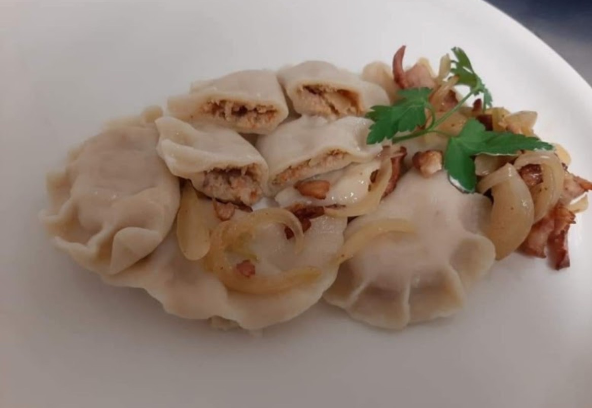 Porcja gotowanych pierogów z mięsnym farszem, część przekrojona, polana podsmażoną cebulą i boczkiem, udekorowana gałązką zielonej pietruszki, na białym talerzu.