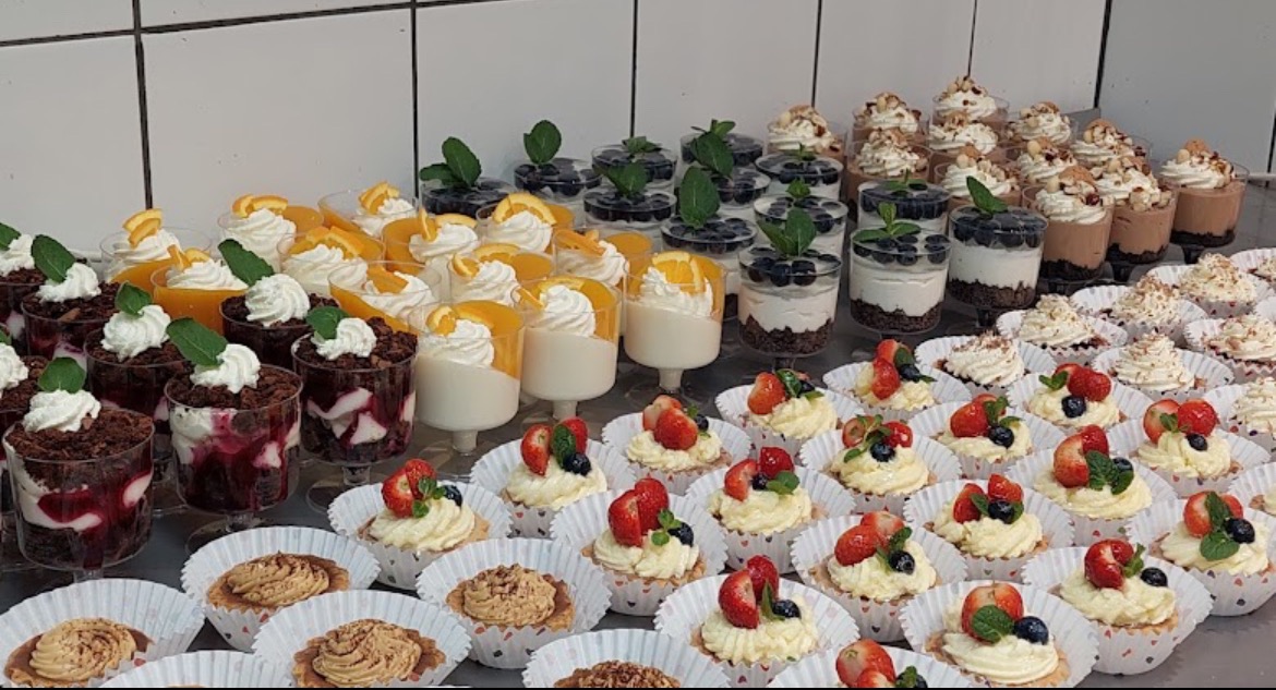 Różnorodny wybór deserów: tartaletki z kremem i truskawkami, musy w przezroczystych pucharkach z owocami i bitą śmietaną, udekorowane listkami mięty.
