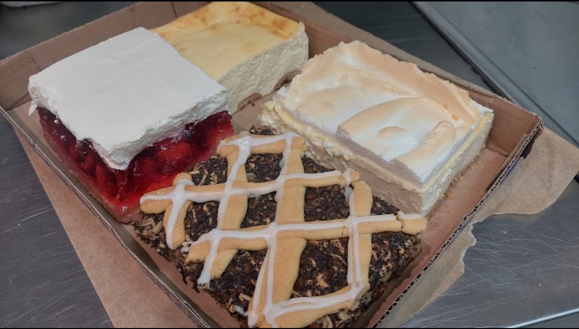 Kartonowe pudełko z czterema różnymi kawałkami ciast: ciasto z galaretką i bitą śmietaną, sernik, ciasto z bezą i ciasto makowe z lukrem w kratkę.