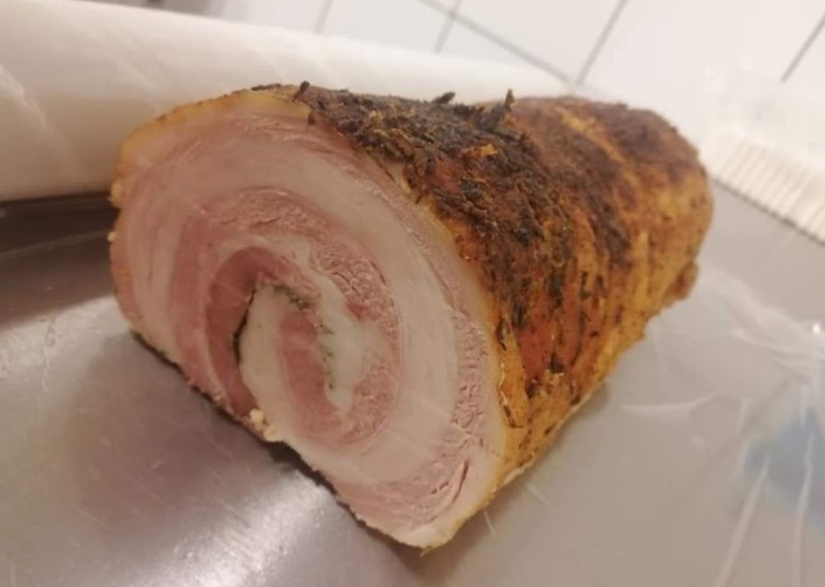 Przekrojona rolada z boczku, z widocznymi warstwami mięsa i tłuszczu, przyprawiona ziołami, leżąca na metalowej powierzchni.