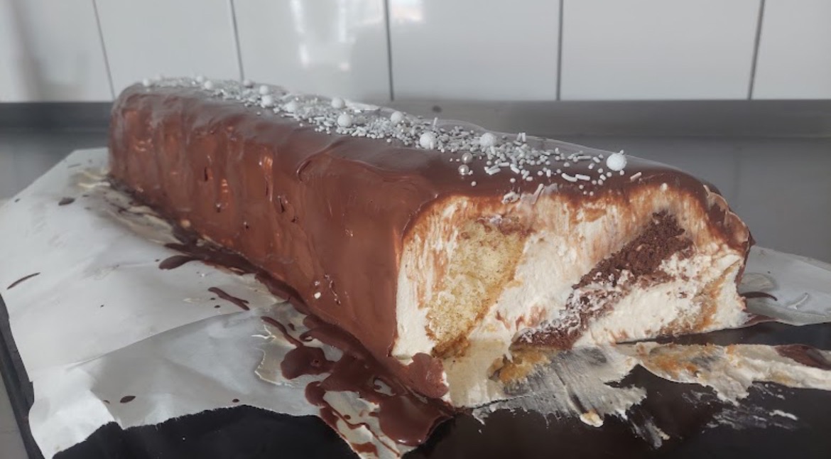 Przekrojony tort prostokątny z kremem i polewą czekoladową, posypany srebrnymi kuleczkami, na papierze do pieczenia.