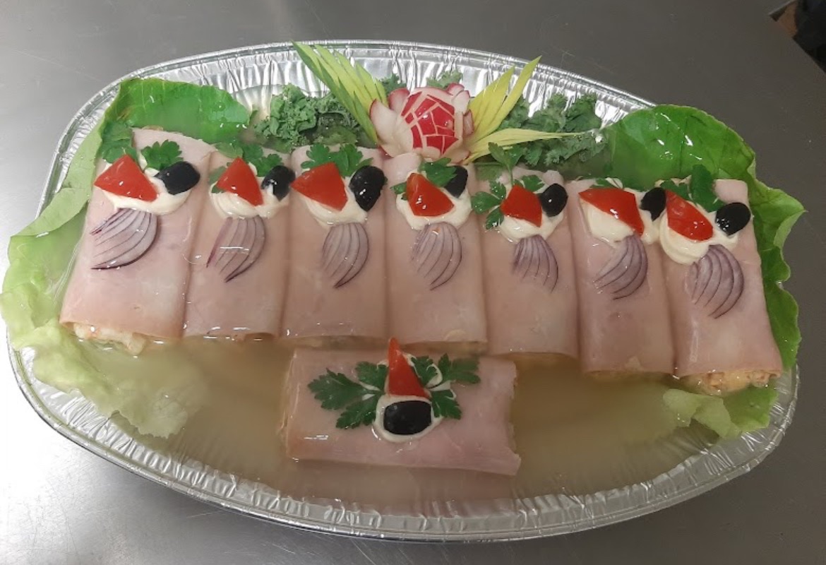 Dekoracyjna galaretka z szynką, udekorowana plasterkami papryki, oliwkami, kleksem majonezu i piórami czerwonej cebuli, podana na tacy z aluminium, otoczonej liśćmi sałaty i rzeźbami z warzyw.