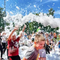 Uśmiechnięty chłopiec w szarej koszulce bawi się w pianie podczas imprezy plenerowej, w tle widoczni inni uczestnicy i zieleń drzew.