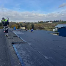 Praca na dachu płaskim: robotnicy układają papę termozgrzewalną, widoczne butle z gazem, narzędzia i fragmenty krajobrazu z zabudowaniami w tle.