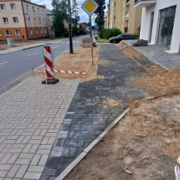 Układanie nowej kostki brukowej na chodniku wzdłuż ulicy, widoczne bariery ochronne i znak drogowy.