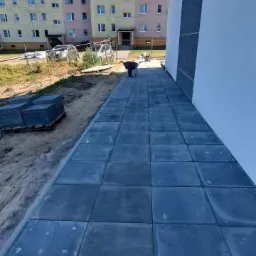 Ułożona kostka brukowa wzdłuż ściany budynku wielorodzinnego, widoczny wózek budowlany i sterta kostki brukowej przed budynkiem.