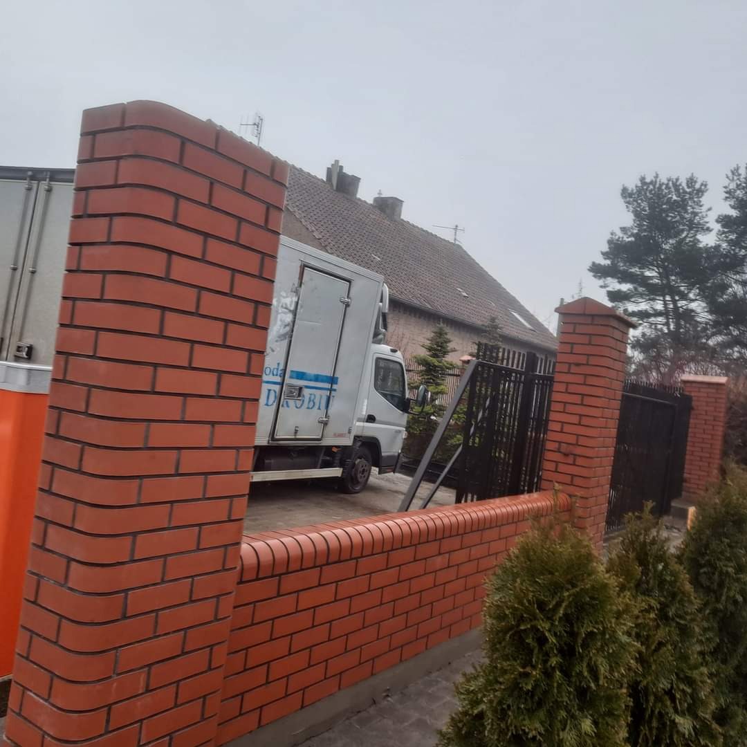 Nowo wybudowany ceglany płot z filarami, z widocznym samochodem dostawczym w tle, na tle domu z ciemnym dachem w pochmurny dzień.
