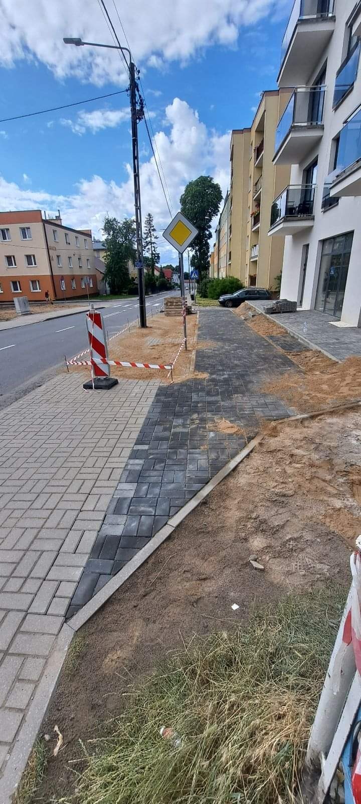 Układanie nowej kostki brukowej na chodniku wzdłuż ulicy, widoczne bariery ochronne i znak drogowy.