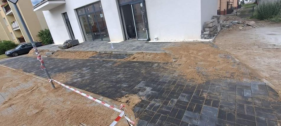 Układanie kostki brukowej przed budynkiem z dużymi oknami, widoczny piasek i taśma ostrzegawcza, kostka ułożona we wzór jodełki.