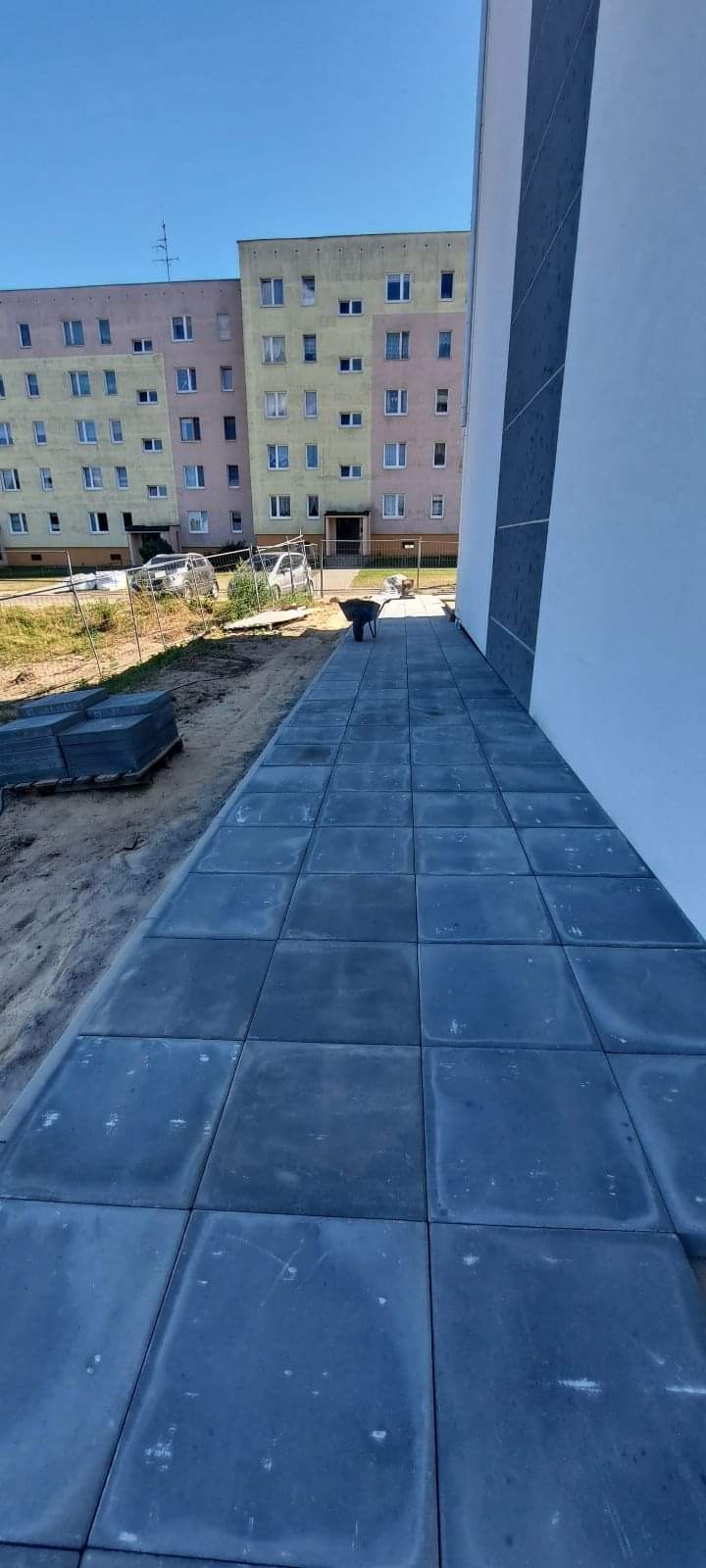 Ułożona kostka brukowa wzdłuż ściany budynku wielorodzinnego, widoczny wózek budowlany i sterta kostki brukowej przed budynkiem.