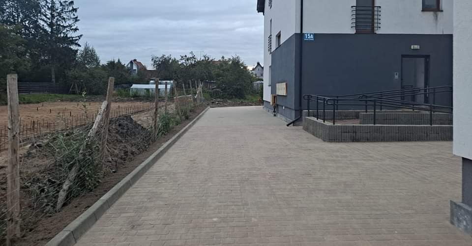 Nowoczesny budynek z szarą elewacją i numerem 15A, widoczny podjazd wyłożony kostką brukową i teren przygotowany pod zagospodarowanie zielenią.