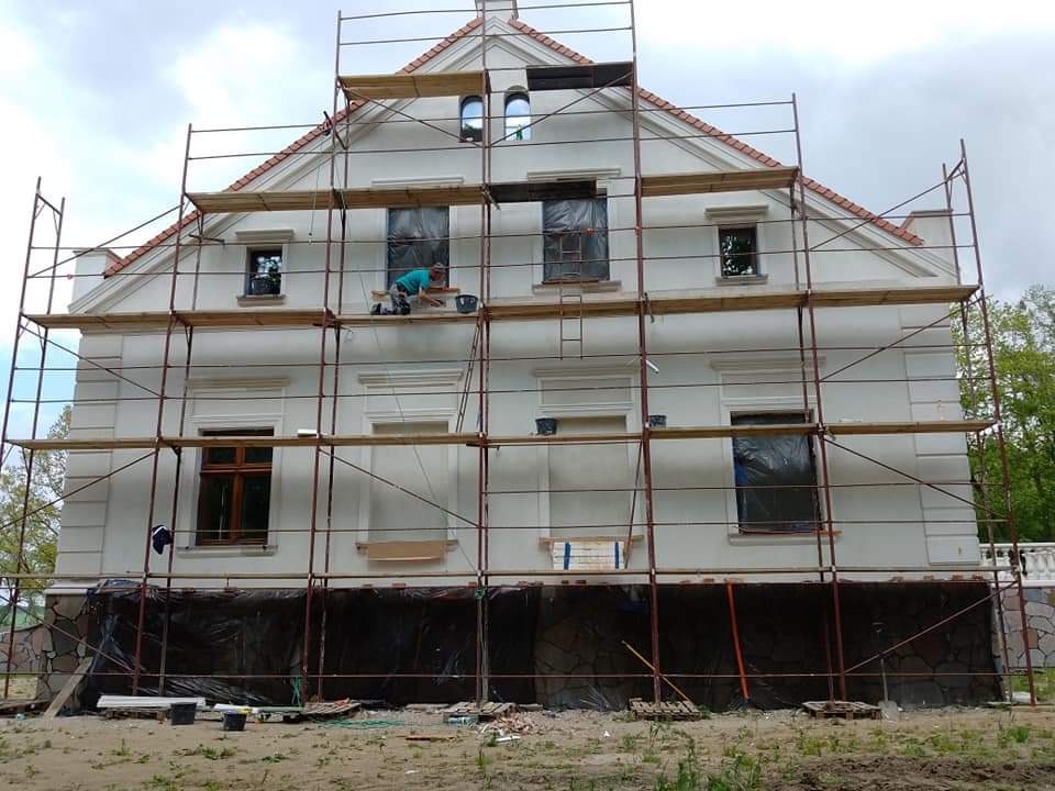 Prace elewacyjne przy dwukondygnacyjnym domu z jasną elewacją, widoczne rusztowania i pracownik w trakcie wykonywania poprawek, dolna część elewacji zabezpieczona folią.