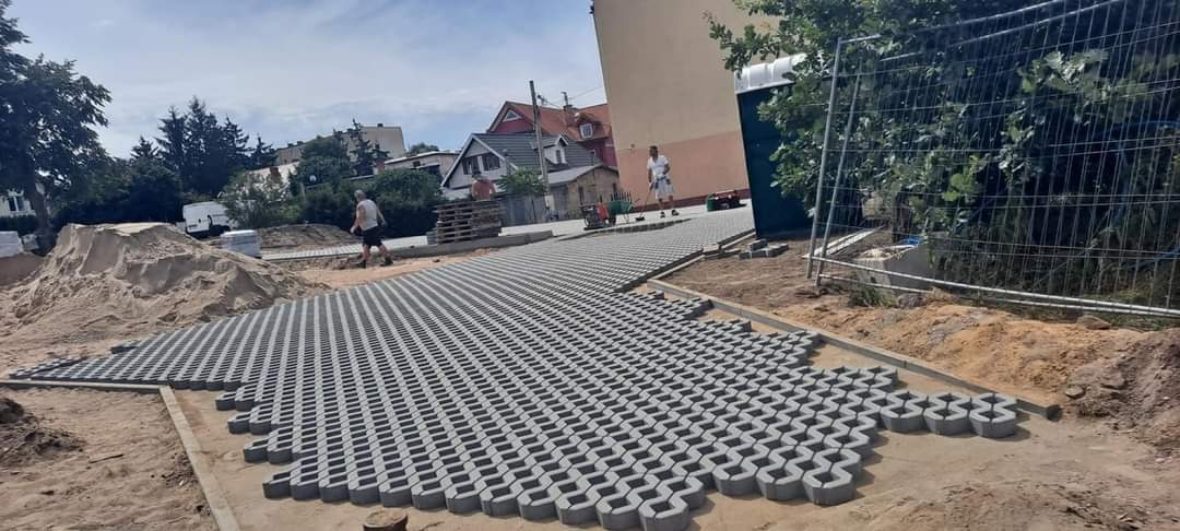 Układanie ażurowych płyt betonowych na piaskowym podłożu, widoczni pracownicy, teren budowy z przenośną toaletą w tle.