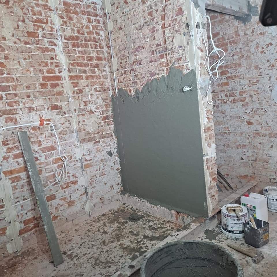 Fragment ściany z czerwonej cegły z nałożoną szarą warstwą świeżej zaprawy, widoczne przewody elektryczne, wiadro z zaprawą i narzędzia murarskie.