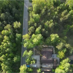 Zdjęcie z drona przedstawia dom z brązowym dachem otoczony gęstym lasem, obok domu widoczny mały staw z dwoma leżakami i stolikiem, a w pobliżu biegnie asfaltowa droga z dwoma samochodami.