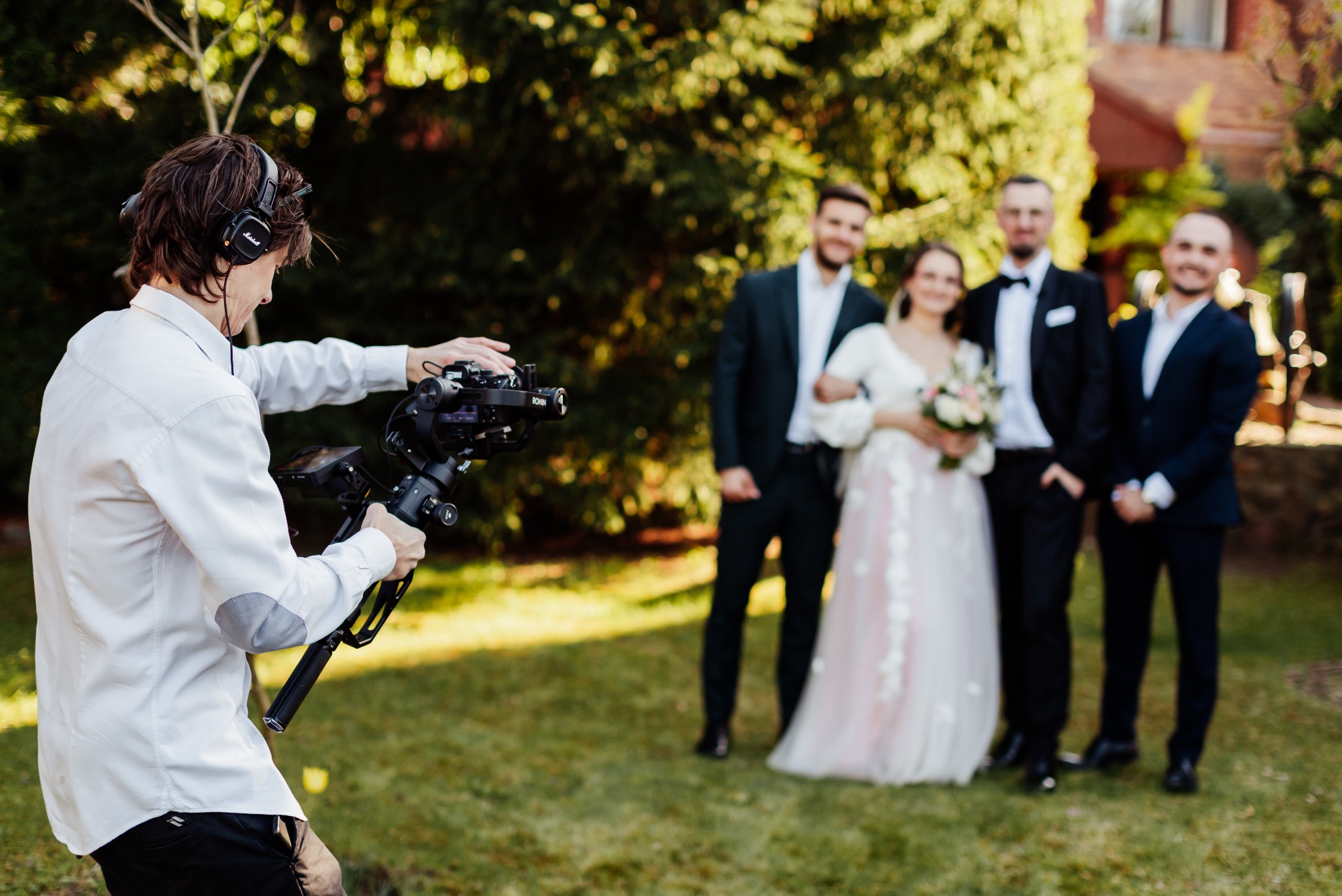 Kamerzysta z gimbalem filmuje parę młodą i gości weselnych w plenerze. Widok zza operatora, na tle zieleni i budynku.
