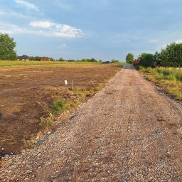 BS-KOP ZMECHANIZOWANE ROBOTY ZIEMNE Bartłomiej Snoch - Droga gruntowa utwardzona kruszywem w Miedzianej Górze, obok przygotowany teren pod budowę, w tle zabudowa jednorodzinna i cień dwóch osób.