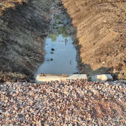 Wykopany rów wypełniony wodą, z betonowymi elementami i warstwą żwiru na pierwszym planie, widoczne ślady prac ziemnych na brzegach rowu.