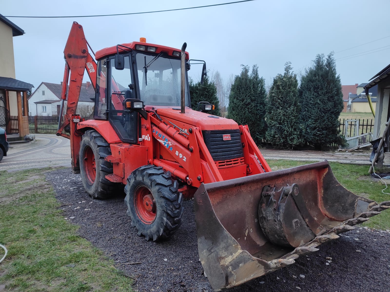 Czerwona koparko-ładowarka FAI Syntesir 892 zaparkowana na żwirowym podłożu, z widocznymi śladami użytkowania na łyżce i ogólnym zabrudzeniem, na tle posesji z tujami i domem.