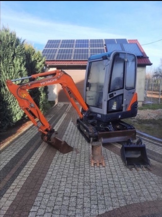 Pomarańczowo-szara minikoparka Hitachi na kostce brukowej, z domem z panelami słonecznymi w tle.