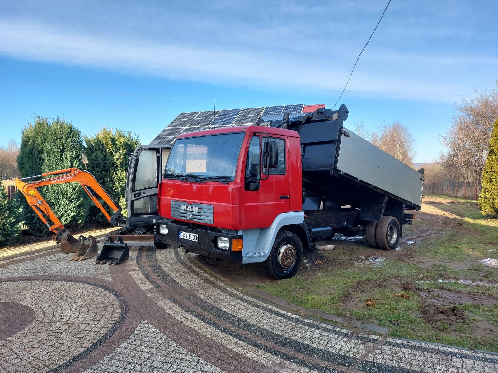 Czerwona wywrotka MAN z podniesioną skrzynią ładunkową stoi na trawie obok pomarańczowej minikoparki Hitachi na kostce brukowej, w tle panele słoneczne na dachu budynku.