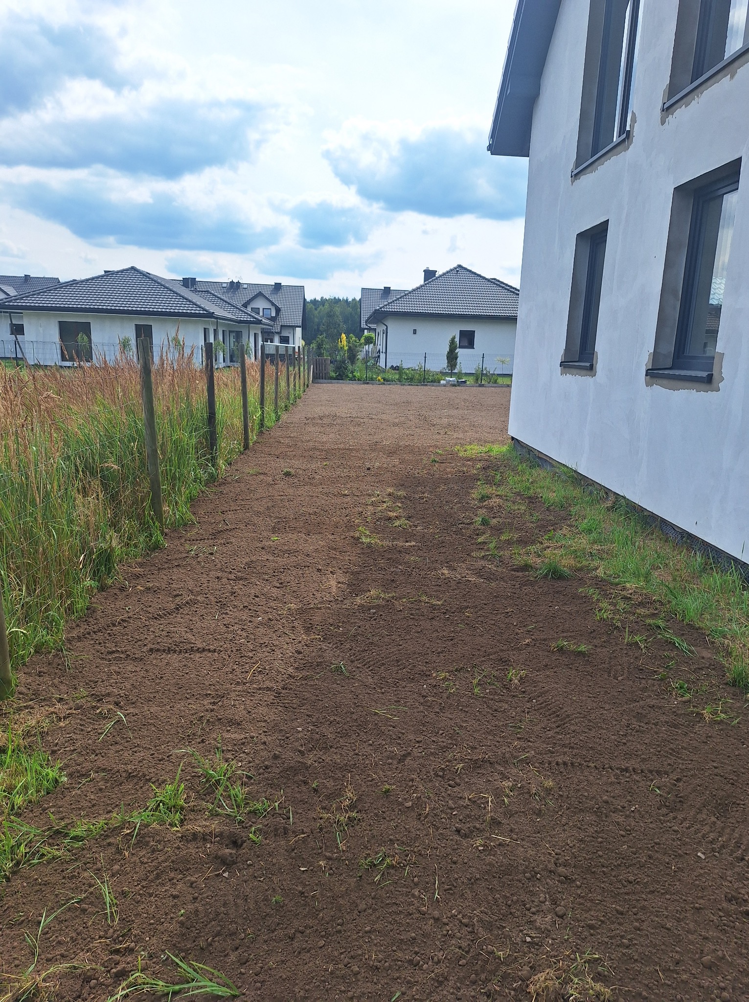 Przygotowany teren pod trawnik z widocznymi śladami po maszynach, obok ogrodzenie z siatki i budynek w stanie surowym otwartym.