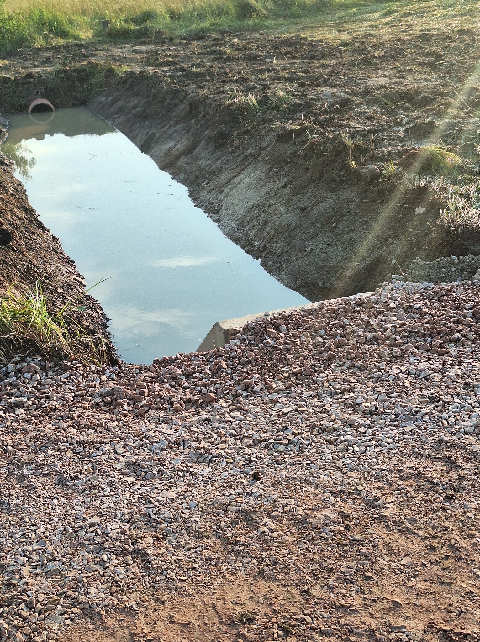 Wykop wypełniony wodą z widoczną rurą drenażową, obok nasyp z kamieni, w tle trawa i ziemia.