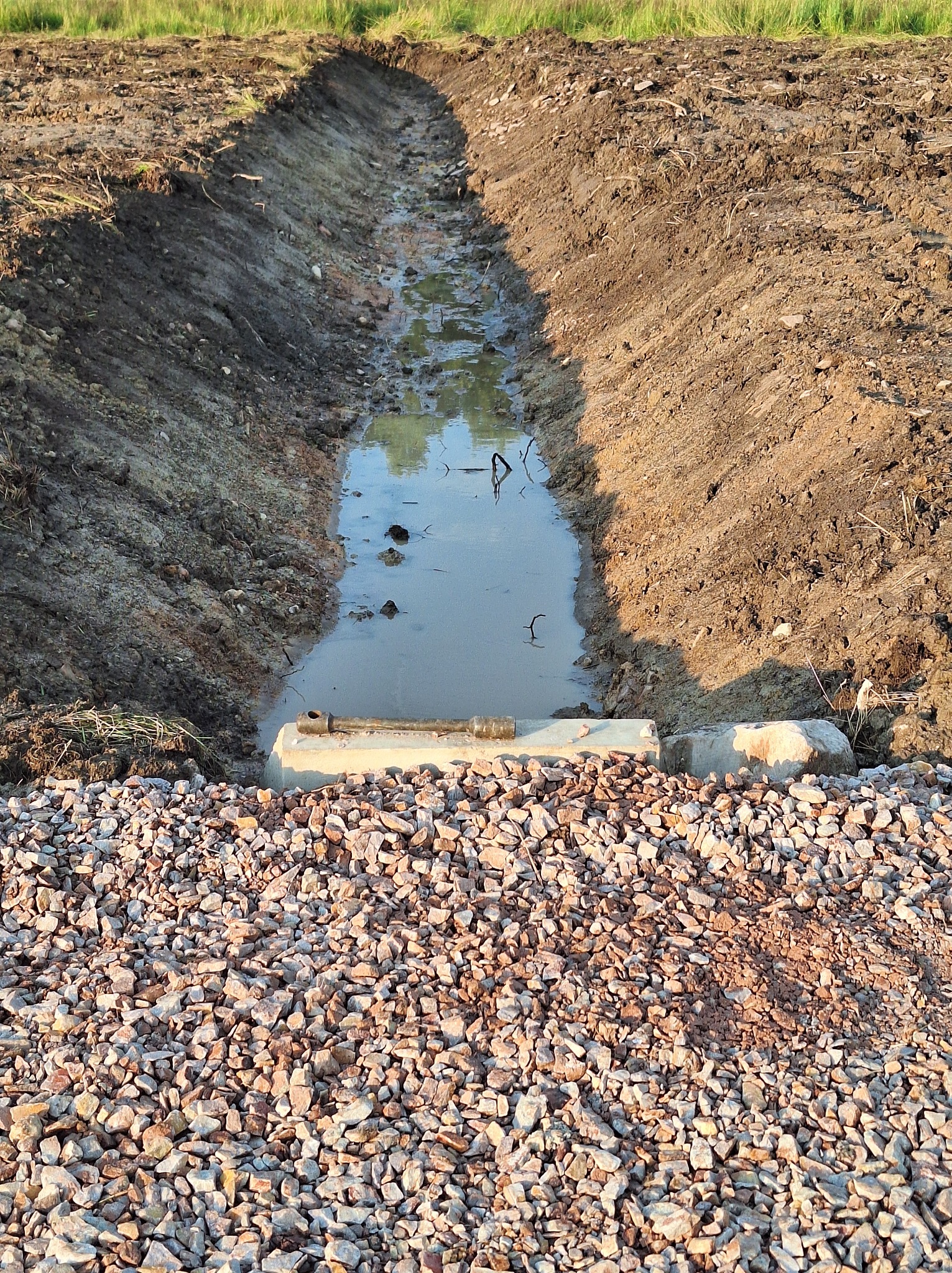 Wykopany rów wypełniony wodą, z betonowymi elementami i warstwą żwiru na pierwszym planie, widoczne ślady prac ziemnych na brzegach rowu.