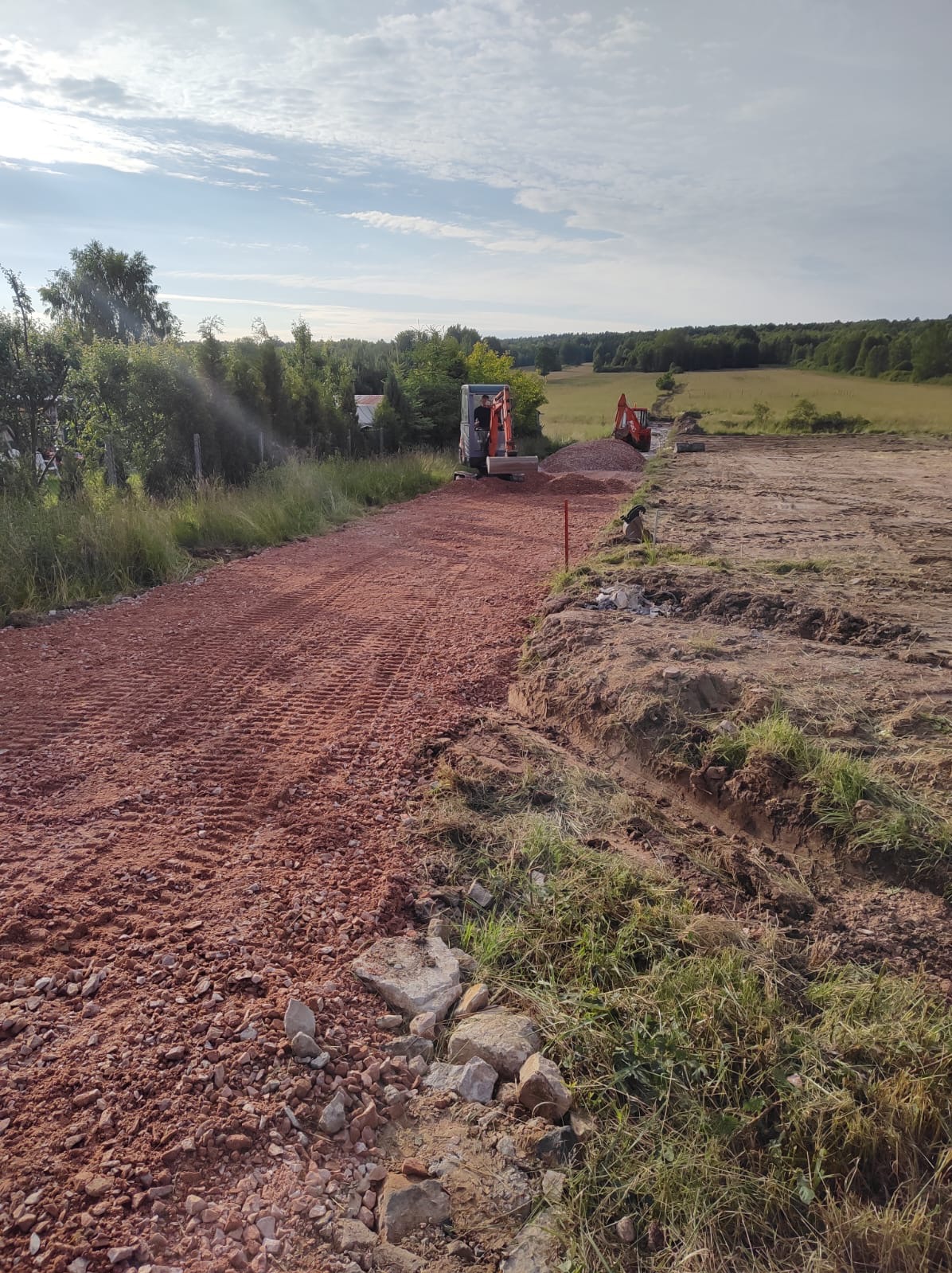 Dwa minikoparki pracują na placu budowy, wyrównując czerwoną, kamienistą nawierzchnię drogi gruntowej; w tle widoczne drzewa i pole.