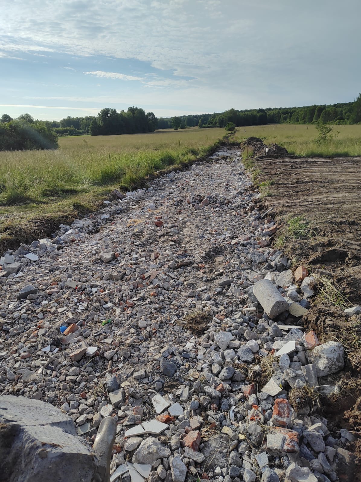 Ścieżka z gruzu i kamieni prowadząca przez łąkę w kierunku lasu, po prawej stronie widoczny fragment przygotowanego gruntu.