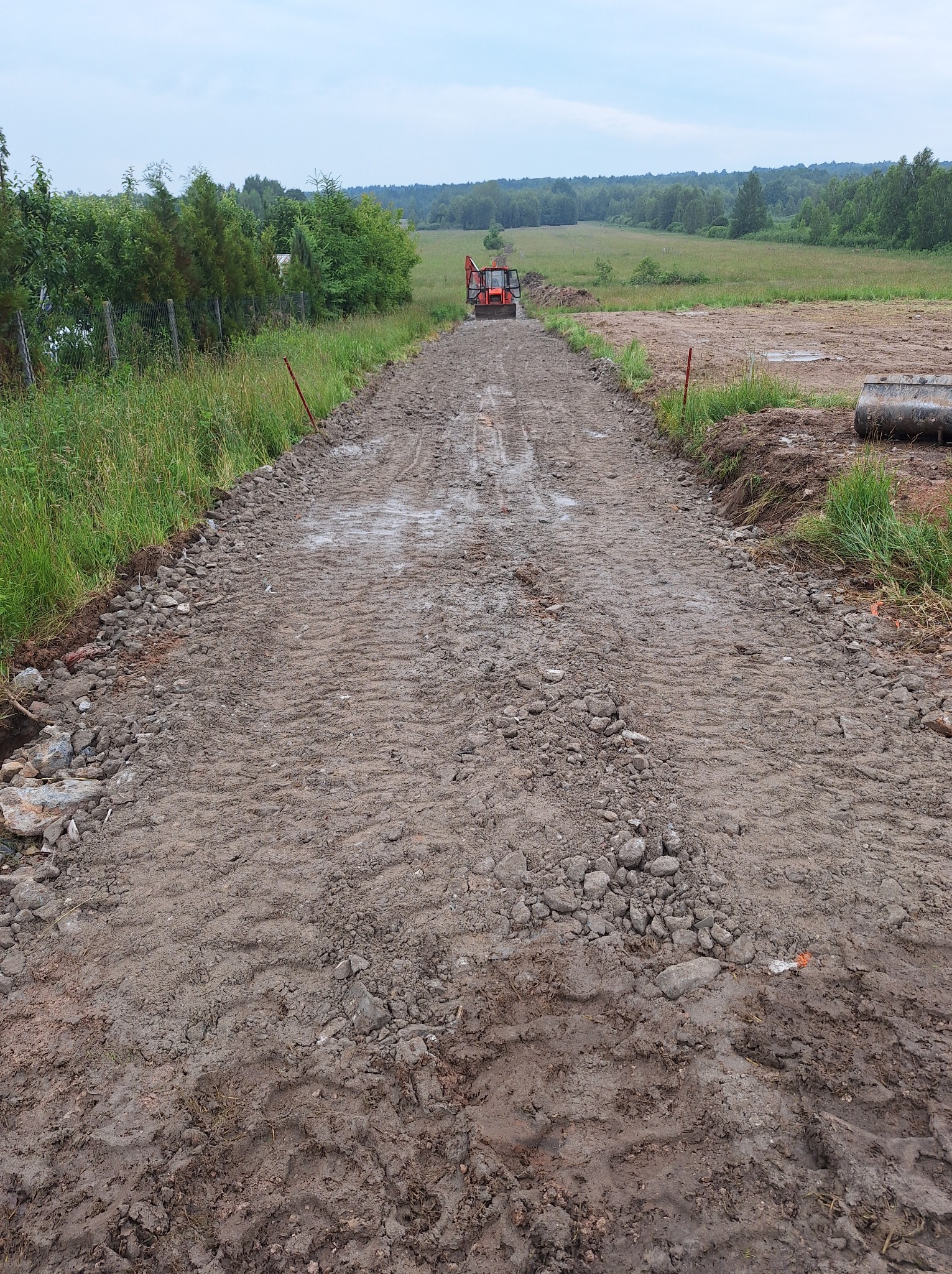 Świeżo wyrównana, błotnista droga gruntowa z widocznymi śladami opon ciągnika w oddali, obrzeża obsypane kamieniami, w tle zielone pola i drzewa.