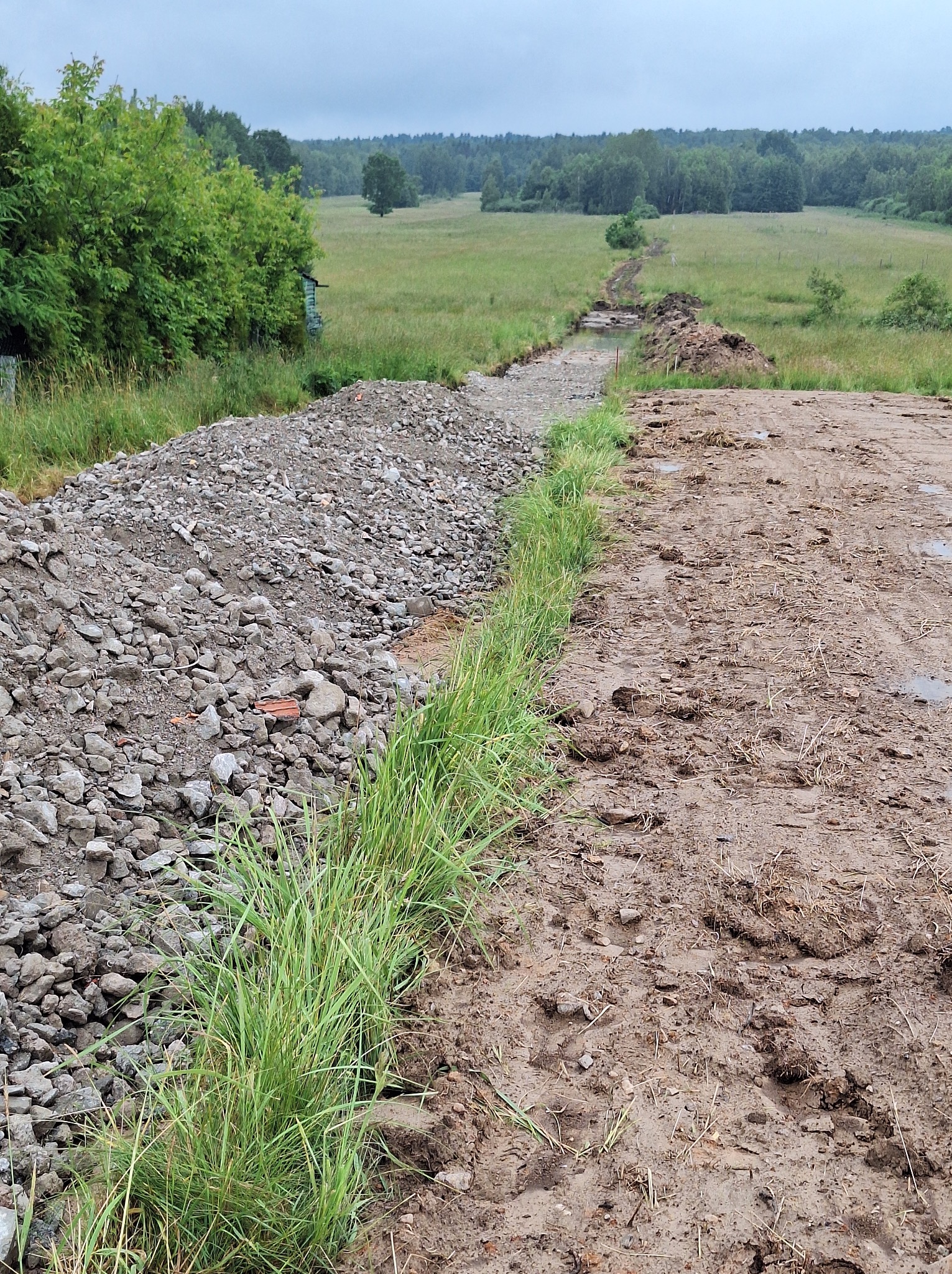 Przygotowanie podłoża pod drogę na działce: hałda kamienia oddzielona pasem trawy od wytyczonego, błotnistego koryta drogi, w tle zielone łąki i las.