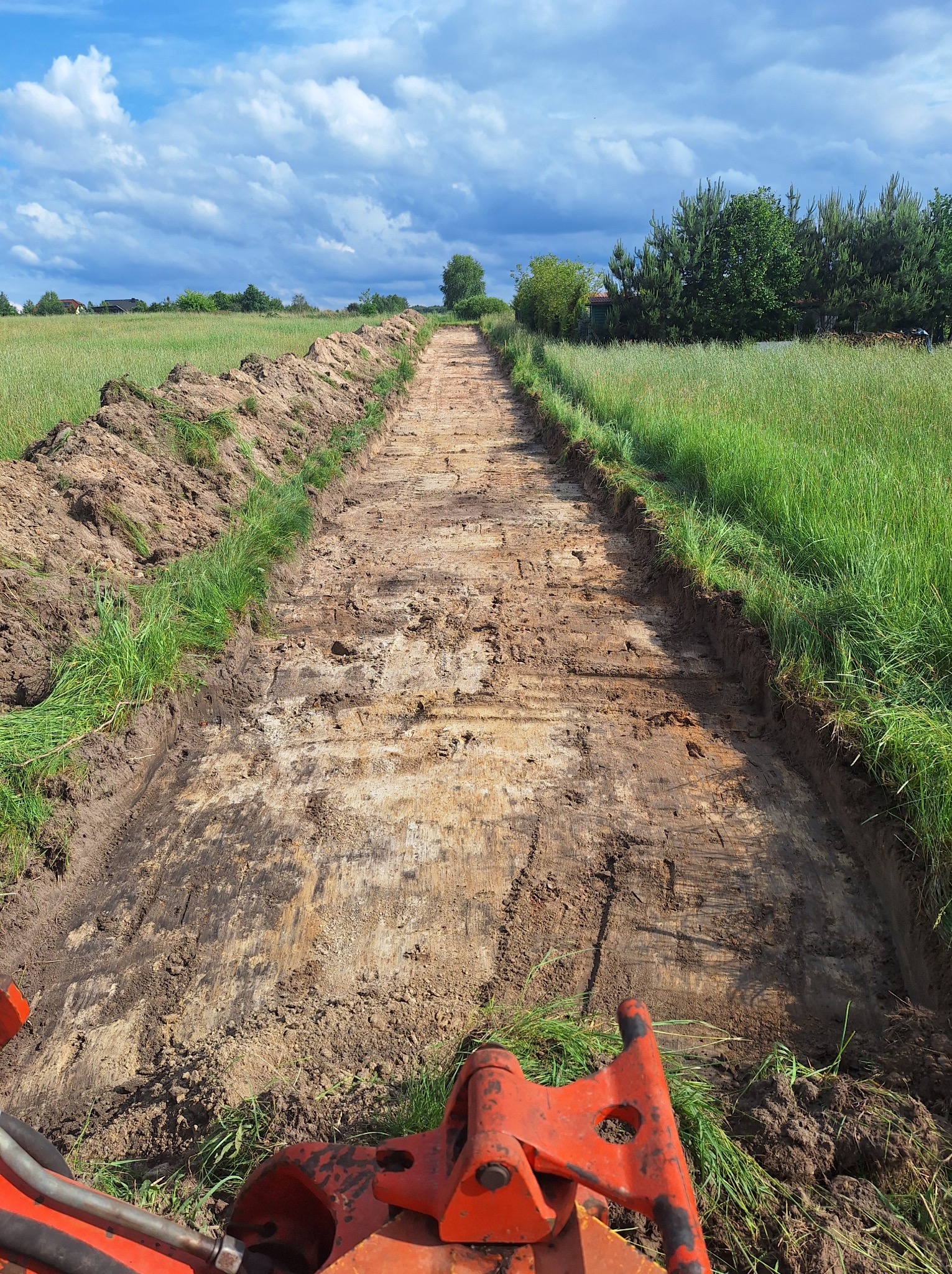 Wąski pas ziemi wykopany w polu, widok z perspektywy operatora maszyny budowlanej, z widocznymi elementami pomarańczowego ramienia koparki na pierwszym planie.