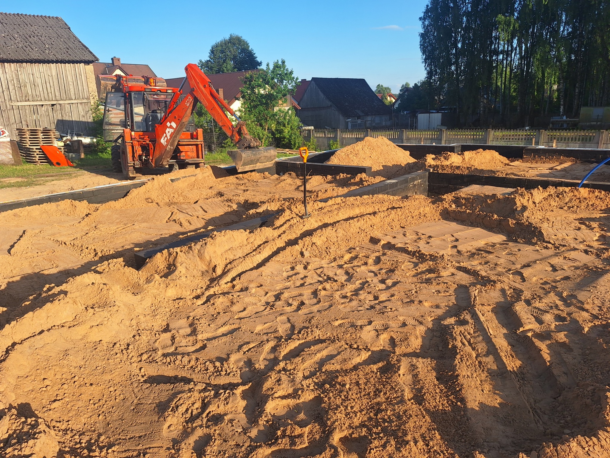 Czerwona koparko-ładowarka FAI podczas prac ziemnych na placu budowy, widoczne wykopane fundamenty, piasek i narzędzia ręczne.