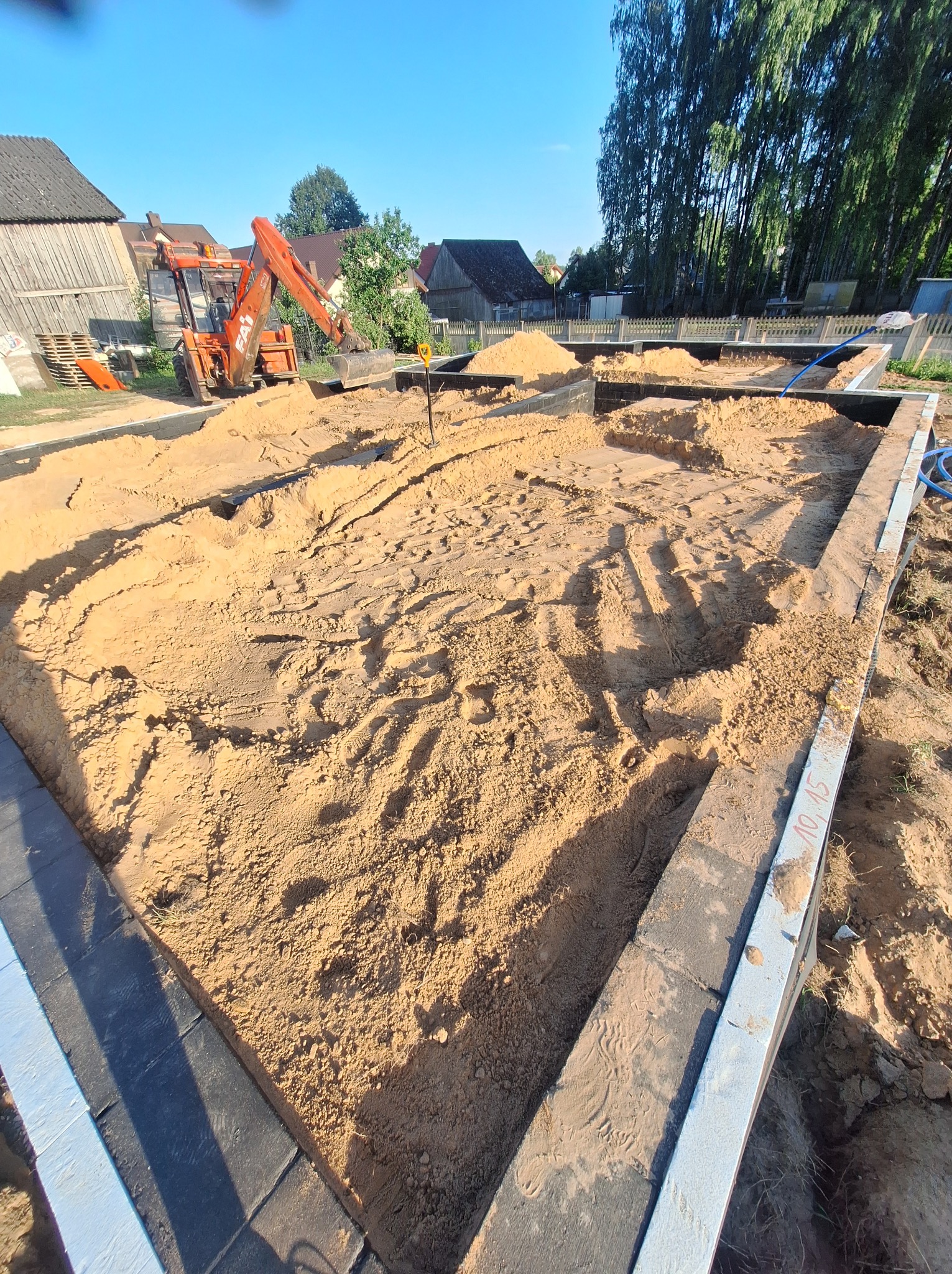Przygotowanie terenu pod fundamenty domu jednorodzinnego, widoczny wykop wypełniony piaskiem, obrys fundamentów wyznaczony przez betonowe bloki, w tle pomarańczowa koparka.
