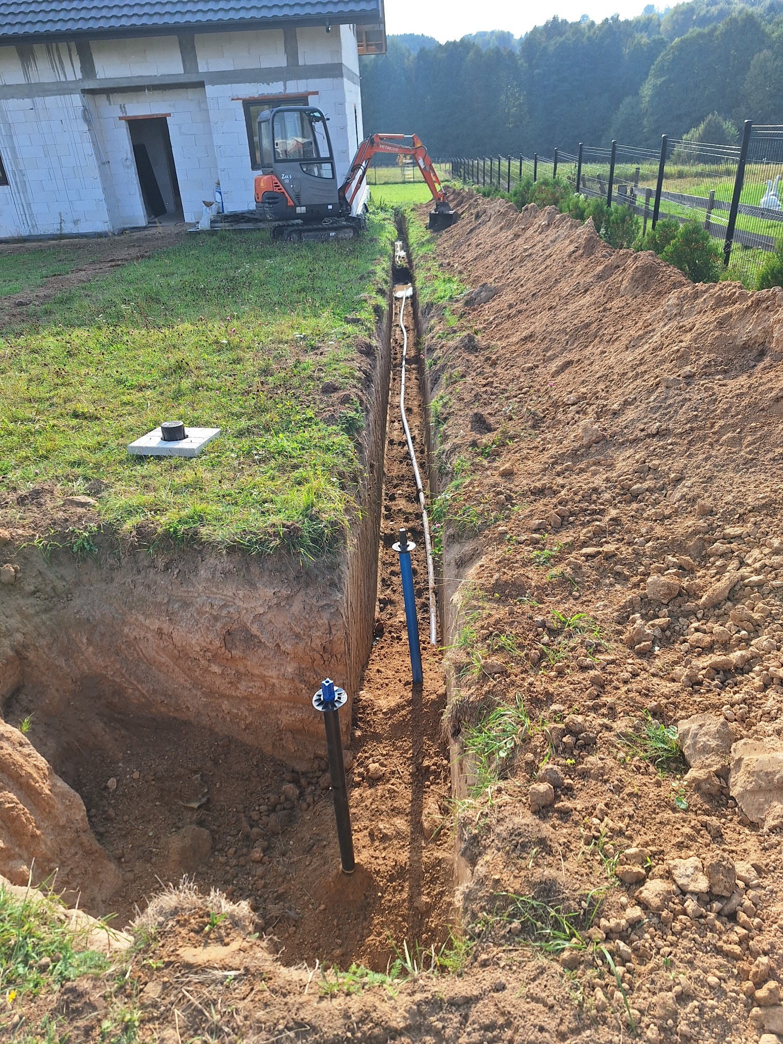 Długi, głęboki wykop w ziemi z widocznymi rurami i przewodami instalacyjnymi, mała koparka stoi obok wykopu na tle budynku w stanie surowym otwartym i zieleni.