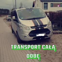 Srebrny bus Ford Transit z czarno-pomarańczowymi pasami na masce i dachu, zaparkowany na chodniku z kostki, widoczny numer telefonu kontaktowego.