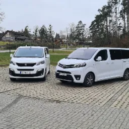 Dwa białe busy: Peugeot i Toyota, zaparkowane na betonowym placu z widocznym budynkiem w stylu rustykalnym w tle.