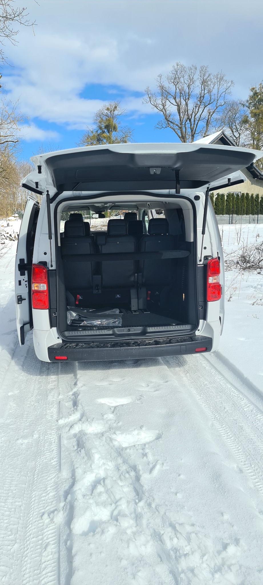 Otwarty bagażnik białego samochodu osobowego z trzema rzędami siedzeń, widok z tyłu na ośnieżoną drogę i zimowy krajobraz.
