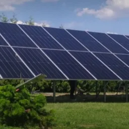 Rozległa instalacja fotowoltaiczna na metalowej konstrukcji, widoczna na tle zielonego trawnika i drzew, z niebieskim niebem w tle.