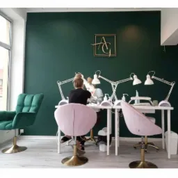 Salon kosmetyczny z ciemnozieloną ścianą, złotym logo i dwoma stanowiskami do manicure z białymi biurkami, lampami i różowymi krzesłami, widok z tyłu na dwie osoby wykonujące zabieg.