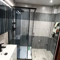 Nowoczesna łazienka z kabiną prysznicową typu walk-in, szare płytki na ścianach i podłodze imitujące drewno, biała umywalka z czarną armaturą, oświetlenie punktowe.