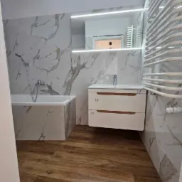 Nowoczesna łazienka po remoncie: drewnopodobna podłoga, białe marmurowe płytki, podświetlane lustro, biała szafka z drewnianymi akcentami, grzejnik drabinkowy.